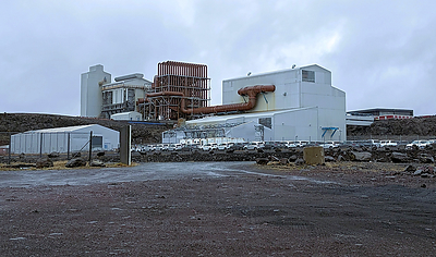 Existing large scale industrial buildings and furnace hall at the Helguvík industrial site in southwest Iceland, illustrating the type of brownfield infrastructure that could potentially be refurbished for future low impact processing activities. The 8,376 m² industrial premises is a former furnace building which previously housed the silicon plant. The building has 5 floors with a lift shaft and 12.5–32.5 ton bridge cranes. The ground floor footprint is about 3,250 m², and each intermediate floor is about 1,430 m² of reinforced concrete slab. The building is on a concrete foundation, built of a steel frame and clad with corrugated sheet metal. The former furnace is a 33 MW electric arc furnace with various possibilities; its internal volume can be reduced, and it can be used for any operation requiring a process temperature of around 1,500 degrees or higher. The furnace building has very strong incoming power (33 kV stepped down to 400 V), glass fiber network, compressed air, four freshwater boreholes, and substantial infrastructure that can be used for almost any industrial production. Photo from Greenland Mines Ltd site visit at Helguvik.