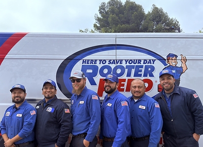 Rooter Hero service technicians stand together in front of a branded company service van, wearing blue uniforms with American flag patches and company hats. The group photo highlights the team ahead of the Fourth of July holiday service period and reflects Rooter Hero’s 24/7 availability and community presence.