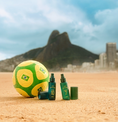 Sol de Janeiro Cheiroso Number 10 and Number 7 cologne mist bottles in beach sand with a Brazil soccer ball and Rio skyline