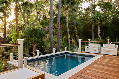 A raised wood deck with white lounge chairs surrounds a rectangular saltwater pool tucked into a canopy of palms and pines.