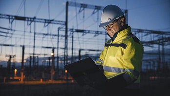Electrical utility worker using a laptop at a power substation at dusk.