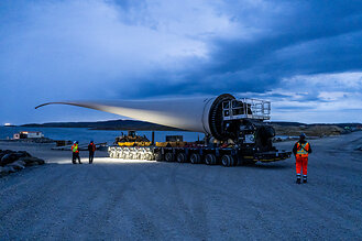 Wind turbine blade being transported on a trailer along a gravel road near the shoreline at dusk. Workers and support vehicles surround the transport under a cloudy evening sky.