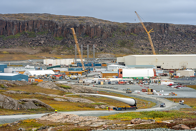 Industrial construction site in a rocky Arctic landscape, with cranes, buildings and equipment beneath a cliff. In the foreground, a wind turbine blade is transported along a gravel road under an overcast sky.