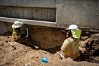 Two Baker Electric workers performing underground conduit work beside a concrete wall.