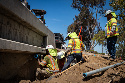 Baker Electric crews installing conduit alongside a concrete freeway structure.