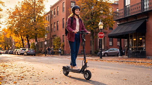 A rider on an Ausom electric scooter navigates a city street lined with autumn foliage during the brand's seasonal sale campaign.