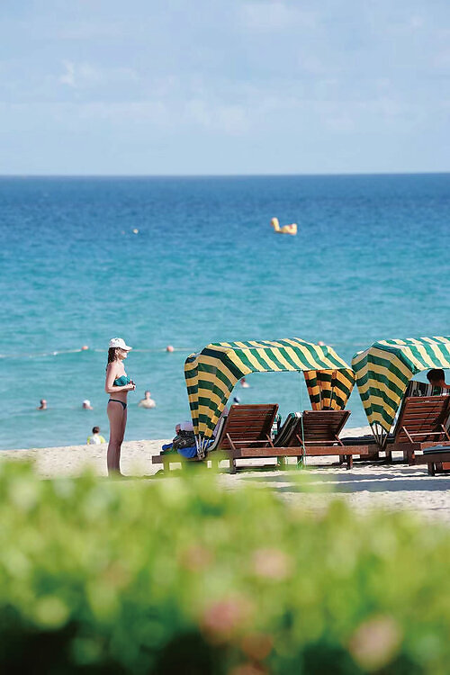 Guests relax on beach loungers under striped canopies at Sanya Marriott Yalong Bay Resort & Spa with the South China Sea in the background. This image opens in the lightbox